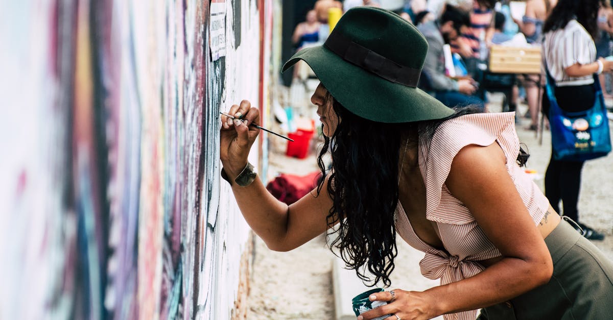 Young woman wearing hat painting a mural on an urban street wall during the day.